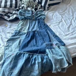 Blå patchwork klänning - Säljer en superfin blå patchwork klänning med olika nyanser av blått. Klänningen har en bohemisk stil med smala axelband och en smockad rygg för bättre passform. Den är dekorerad med små knappar framtill och har en volangdetalj längst ner. Perfekt för sommardagar eller en avslappnad fest!
