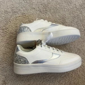 Vita sneakers med glittriga detaljer - Säljer ett par snygga vita sneakers med glittriga silverdetaljer på hälen och sidan. Skorna har en klassisk snörning och en bekväm vit sula. Bara testade så inprincip oanvända. Hör av dig vid frågor eller funderingar 
