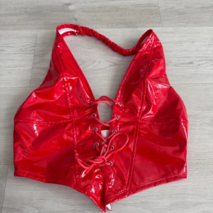 Röd glansig korsettopp - Säljer en supercool röd glansig korsettopp med snörning framtill. Toppen är ärmlös och har en djup v-ringning. Perfekt för en festkväll när du vill sticka ut!