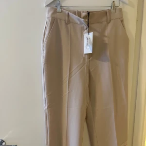 Beige byxor från NA-KD Reborn - Säljer ett par snygga beige byxor från NA-KD Reborn. De är högmidjade och har en croppad stil som ger en modern look. Perfekta för både vardag och fest! Byxorna har en stilren design med knappar och dragkedja framtill.