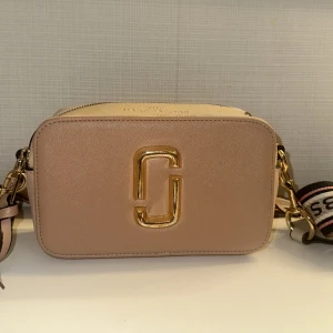 Beige axelväska från Marc Jacobs - Säljer en superfin rosa axelväska från Marc Jacobs. Väskan har en snygg guldig logga på framsidan och en justerbar axelrem med texten 'THE MARC JACOBS'. Perfekt för både vardag och fest! Köpt för 999 kr och säljer för 450 kr. Ser även real ut.