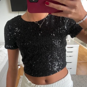 Svart glittrig croppad topp - skitsnygg svart glittrig topp med paljetter. perfekt för nyår och fest! Toppen har en öppen rygg som knyts längst ner, vilket ger en extra snygg detalj. endast använd 1 gång så precis som ny!