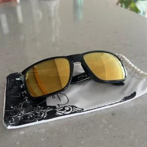 Svarta solglasögon med guldfärgade linser - Säljer ett par snygga svarta Oakley solglasögon med spegelblanka guldfärgade linser. De har en cool och sportig design. Perfekta för soliga dagar och ger en riktigt skarp look. Kommer med en vit och svart förvaringspåse. Sprickor i glaset runt om. 
