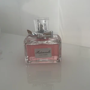 Mademoiselle Pour Femme parfym - Säljer en elegant parfymflaska med rosa doftvätska. Flaskan har en stilren design med en silverfärgad rosett på korken. Perfekt för den som älskar en feminin och sofistikerad doft. Passar både till vardags och fest!