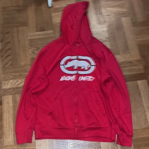 Röd hoodie från Ecko Unltd - Säljer en cool röd hoodie från Ecko Unltd med dragkedja och stort tryck av deras ikoniska logga på framsidan. Perfekt för en avslappnad stil och passar bra till både höst och vår. Den har en skön passform och är i bra skick. Perfekt för streetwear-älskare! 🔥