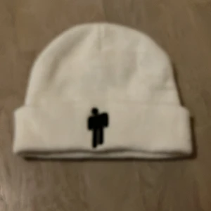 Billie eilish mössa vit - Säljer en stilren vit mössa med en cool svart broderad logga framtill. Perfekt för att hålla sig varm under kyliga dagar. Mössan är i bra skick och passar de flesta tack vare sin flexibla passform.