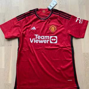 Säljer en ny röd Manchester United fotbollströja från Adidas. Tröjan har korta ärmar och är i perfekt skick med klubbens emblem och sponsorlogga på framsidan. Den är tillverkad i ett lätt och andningsbart material, perfekt för både träning och matchdagar. 