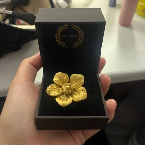 Guldfärgad blomring från Olympia - Säljer en superfin guldfärgad ring från Olympia, formad som poppy blomma. Använt endast 2 gånger, storlek S (16,5mm). Perfekt för att ge en extra touch till din outfit, oavsett om det är till vardags eller fest. Kommer i en snygg presentask.