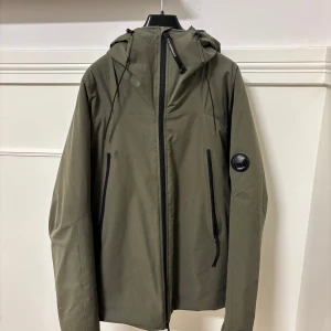 Olivgrön jacka från C.P. Company - Säljer en olivgrön jacka från C.P. Company i mycket bra skick. Jackan är tillverkad i stretchig polyester med WR-beläggning och har en dragkedja framtill. Den har en huva och långa ärmar, perfekt för höst och vår men fungerar även att använda som vinterjacka. Märket är synligt på ärmen och insidan har Pro-Tek och Primaloft detaljer. Passar S och M.