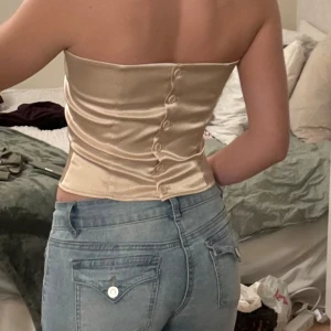 Beige halterneck korsett - Snygg beige halterneck korsett i sidenliknande material. Toppen har knappar längs ryggen och en stilren design som passar perfekt för festliga tillfällen. Den är tight och ger en elegant look.🤍🩷Storlek M, men skulle säga S!