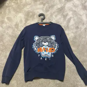 Säljer en snygg mörkblå sweatshirt från Kenzo med ett coolt tigertryck i orange och blått på framsidan. Tröjan har långa ärmar och är i mycket bra skick. Perfekt för både vardag och fest!