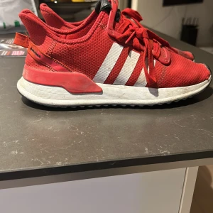 Röda Adidas sneakers - Säljer ett par röda Adidas sneakers i bra skick. De har en snygg design med vita ränder på sidan och är perfekta för både vardag och träning. Skorna har snörning och en bekväm sula som ger bra stöd. Passar perfekt för dig som vill ha en sportig och stilren look.