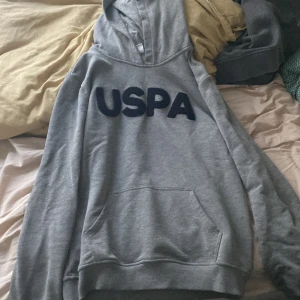 Grå hoodie från USPA - Säljer en grå hoodie från USPA i bra skick. Den har en stor, mysig huva och en praktisk magficka. Perfekt för kyliga dagar eller bara för att chilla hemma. Stor USPA-logga på framsidan ger en sportig look. Passar både till vardags och avslappnade tillfällen.