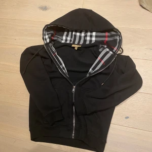 Svart Burberry Zipup - Säljer en snygg svart hoodie från Burberry i storlek S. Den har ett klassiskt rutigt foder i huvan med grå, vita och röda detaljer. Dragkedja framtill och broderad logga på bröstet. Perfekt för höst och vinter!