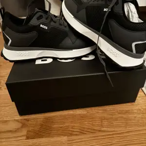 Säljer ett par snygga svarta sneakers från Boss i nytt skick. De har en stilren design med vita detaljer och snörning. Perfekta för både vardag och fest! Kommer i originalkartong. Storlek 43