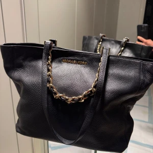 Svart skinnväska från Michael Kors - Säljer en snygg svart skinnväska i lammskinn från Michael Kors. Väskan är i nyskick och har kedjedetaljer på handtagen i guld och flera fack inuti för smart förvaring. Perfekt för både vardag och fest! 🖤 Den är ca 30cm B, 25cm H och 12cm djup