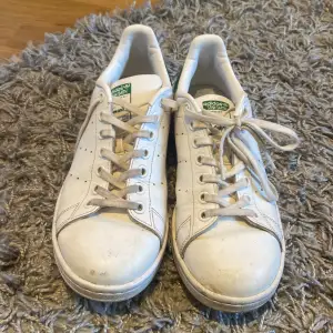 Säljer ett par klassiska Adidas Stan Smith sneakers i vitt med gröna detaljer. Skorna är i bra skick med lite smuts på ovansidan. Perfekta för vardagsbruk och ger en stilren look. De har snörning och är gjorda i syntetmaterial.