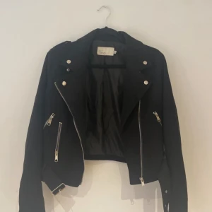 Svart mockajacka från NLY Trend - Säljer en snygg svart mockajacka från NLY Trend i storlek 36. Jackan har coola dragkedjedetaljer och knappar framtill. Perfekt för höst och vår, och ger en edgy look till vilken outfit som helst. Passar både till vardags och fest!