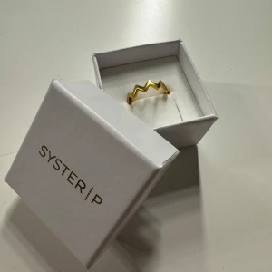 Guldfärgad ring från Syster P - Snygg guldfärgad ring från Syster P med ett unikt, zickzack-mönster. Perfekt för att ge en modern touch till din outfit. Ringen kommer i en elegant vit ask, vilket gör den till en fin presentidé. Passar både till vardags och festliga tillfällen.