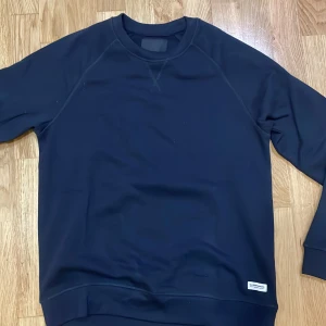 Mörkblå sweatshirt - Säljer en stilren mörkblå sweatshirt i mycket bra skick. Den har en enkel design med långa ärmar och en liten etikett nertill. Perfekt för både vardag och avslappnade tillfällen. Passar bra till jeans eller chinos!