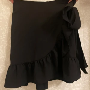 Svart omlottkjol från Vero Moda - Säljer en superfin svart omlottkjol från Vero Moda i storlek XS. Kjolen har en volangdetalj längs nederkanten och knyts i midjan för en perfekt passform. Perfekt för både vardag och fest! 🖤