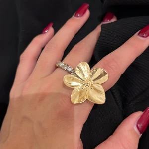 Guldfärgad blomring - Säljer en superfin guldfärgad ring med en stor blomma som detalj. Perfekt för att ge en extra touch till din outfit. Ringen är i mycket bra skick och passar perfekt för festliga tillfällen. Den har en elegant och unik design som verkligen sticker ut.