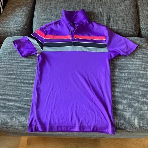 Lila randig pikétröja från Under Armour - Säljer en snygg lila pikétröja från Under Armour med färgglada ränder i rosa, svart och grått över bröstet. Perfekt för både träning och vardag. Tröjan har korta ärmar och en klassisk krage med knappar. Passar perfekt för en sportig look! Pris går alltid att förhandlas!