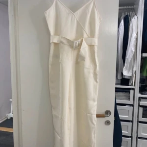 Vit byxdress - Säljer en elegant vit byxdress med omlott-design och bälte i midjan. Perfekt för sommarens fester eller studentfiranden. Byxdressen har en lång passform och är tillverkad i ett mjukt material som ger en fin siluett.