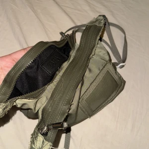 Olivgrön axelväska med dragkedja - Säljer en olivgrön axelväska i syntetmaterial. Väskan har en praktisk dragkedja och justerbar axelrem. Perfekt för vardagsbruk och enkel att matcha med olika outfits. Den har en sliten look som ger den karaktär. KÖPT FRÅN ZARA