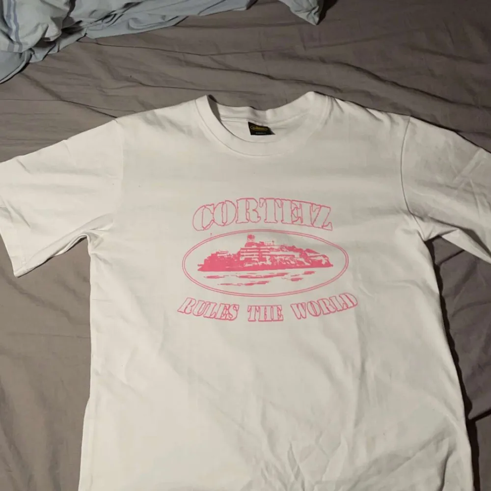 Säljer en vit t-shirt från Cortez i storlek small. Den har ett coolt rosa tryck med texten 'Cortez Rules The World' och en bild i mitten. Perfekt för en avslappnad stil eller som statement piece. T-shirten är i bomull och har korta ärmar.. T-shirts.