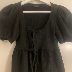 Svart ribbad klänning från Gina Tricot - Säljer en svart ribbad klänning från Gina Tricot i storlek XS. Den har puffiga kortärmar och en knytning framtill som ger en söt detalj. Perfekt för både vardag och festliga tillfällen!