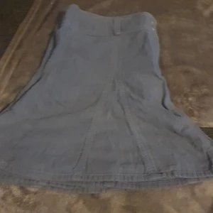 Blå jeanskjol - Säljer en snygg blå jeanskjol i bra skick. Kjolen har en klassisk design med två knappar framtill och bälteshällor. Perfekt för både vardag och festliga tillfällen. Passar bra till en avslappnad stil med sneakers eller klä upp med klackar.