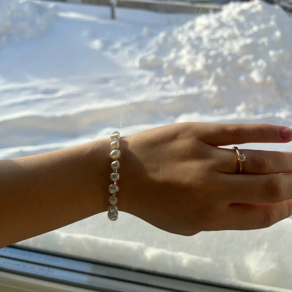 Elegant armband med vita pärlor och guldfärgad låsdetalj. Armbandet har en fin liten hängande dekoration i form av en glittrig insekt. Perfekt för att ge en touch av lyx till din outfit, oavsett om det är för vardag eller fest.. Asusteet.