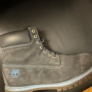 Grå boots från Timberland - Säljer ett par grå boots från Timberland i mocka. De har en robust design med snörning och en bekväm vadderad krage. Perfekta för höst och vinter med sin slitstarka sula. Timberland-loggan syns på sidan av skon. Passar både för stad och natur.