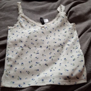 Blommig linne med spetsdetaljer - Söt blommig linne i vitt med små blå blommor och gröna blad. Den har spetsdetaljer vid axelbanden och en liten rosett framtill. Perfekt för sommardagar eller som ett lager under en kofta.