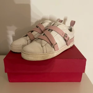 Valentino sneakers med rosa detaljer - Säljer ett par snygga Valentino sneakers i vitt skinn med coola rosa remmar och nitar. De har en unik design med spännen och är perfekta för att ge din outfit en lyxig touch. Skorna är i bra skick och kommer i originalkartong.