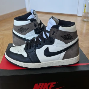 Jordan 1 Mocha - Säljer ett par Nike Air Jordans i färgerna svart, vit och brun. Skorna är i bra skick med lite slitage på sulan. De har en klassisk design med snörning och Nike-loggan på sidan. Perfekta för både vardag och träning. Vid snabb affär 750kr
