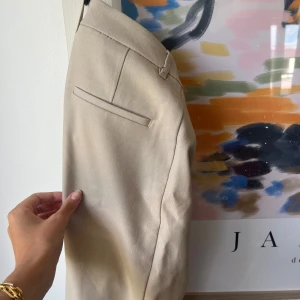 Beige kostymbyxor - Säljer ett par stiliga beige kostymbyxor. De är högmidjade och har en rak passform. Byxorna har en enkel design med en diskret ficka på baksidan och är perfekta för både formella och semi-formella tillfällen. Materialet känns mjukt och bekvämt.