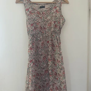 Blommig ärmlös klänning - Säljer en superfin blommig klänning som är perfekt för sommaren! Den är ärmlös och har ett härligt mönster med röda och vita blommor på en beige bakgrund. Klänningen är i normal passform och är gjord av ett mjukt och skönt material, troligtvis bomull eller polyester. 