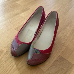 Färgglada ballerinaskor med klack - Säljer ett par unika ballerinaskor med klack i storlek 37. Skorna är i en mix av rött och brunt med färgglada detaljer och mönster. De har en bekväm passform och är perfekta för både vardag och fest. Skorna är i bra skick och redo för en ny ägare!