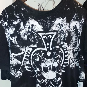 Svart t-shirt med dödskalletryck från Rusty Neal - Säljer en svart t-shirt från Rusty Neal med ett coolt dödskalletryck i vitt. T-shirten har korta ärmar och en normal passform. Perfekt för en rockig stil eller för att sticka ut på festen!