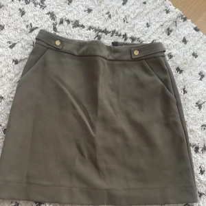 Olivgrön kjol med knappar - Säljer en snygg olivgrön kjol i storlek 38. Kjolen har en enkel och stilren design med två knappar framtill. Perfekt för både vardag och fest! Materialet känns mjukt och bekvämt.