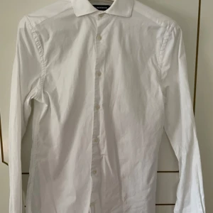 Vit skjorta från J.Lindeberg - Säljer en klassisk vit skjorta från J.Lindeberg. Den har långa ärmar och knappar hela vägen ner. Perfekt för både formella och avslappnade tillfällen. Skjortan är i bra skick och gjord av ett bekvämt material.