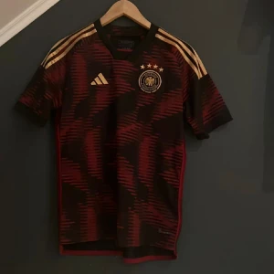 Tysklands fotbollströja! - Säljer en snygg fotbollströja från Adidas i svart och rött med guldiga detaljer. Tröjan har korta ärmar och ett coolt mönster. Perfekt för fotbollsfans som vill ha något unikt! 🌟Tysklands gamla borta ställ!😍