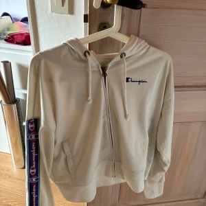 Beige hoodie från Champion - Säljer en snygg vit hoodie från Champion i storlek S. Den har en dragkedja framtill och långa ärmar med coola Champion-loggor längs sidorna. Perfekt för en avslappnad stil och passar bra till både höst och vår. Superbekväm och i bra skick!