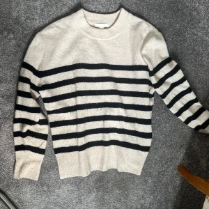 Randig stickad tröja - Säljer en supermysig stickad tröja med svart och beige ränder. Perfekt för kyliga dagar! Den har långa ärmar och en klassisk rund hals. Passar både till vardags och lite finare tillfällen. 🖤🤍