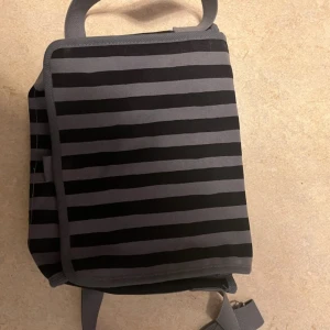 Randig axelväska från Marimekko - Säljer en snygg randig axelväska från Marimekko. Väskan är i svart och grått med ett praktiskt lock och knappstängning. Perfekt för vardagsbruk och har en justerbar axelrem för bekväm passform. Nyskick 