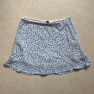 Blommig kortkjol i blått och vitt - Säljer en superfin kortkjol med ett sött blommönster i blått och vitt. Kjolen är perfekt för sommardagar och har en lätt och luftig känsla. Den är i nyskick och passar perfekt till både vardag och fest. Ett måste i garderoben för alla som älskar blommiga mönster!