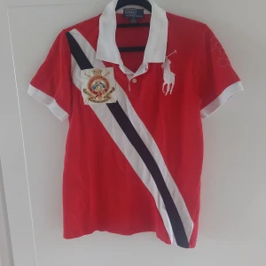 Röd kortärmad pikétröja från Ralph Lauren - Säljer en snygg röd pikétröja från Ralph Lauren i storlek S. Tröjan har en vit och svart diagonal rand och ett stort broderat emblem på bröstet. Den har också en vit krage och nummer 3 broderat på ärmen. Perfekt för en casual och stilren look!