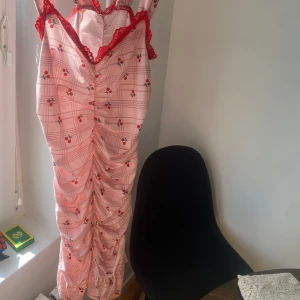 Rosa långklänning med rutor och spetsdetaljer - Säljer en superfin rosa långklänning med rutigt mönster och små söta körsbär. Klänningen har röda spetsdetaljer vid bysten och axelbanden. Perfekt för sommarens fester eller en mysig picknick i parken! 🍒